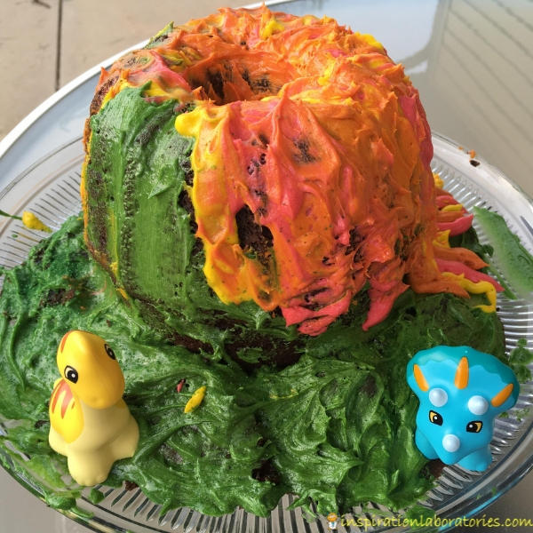 volcano birthday cake decorated with red, orange, yellow, and green buttercream frosting with dinosaur toys