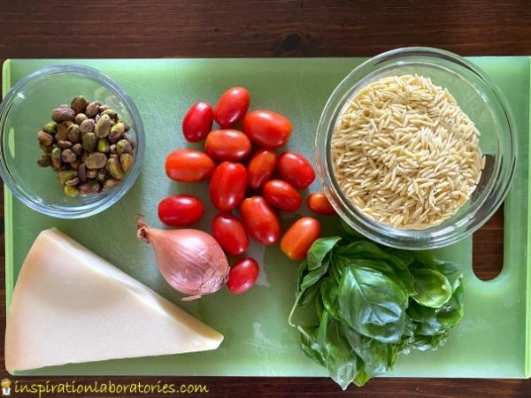 orzo pasta recipe ingredients - orzo, tomatoes, shallots, pistachios, parmesan, and basil on a cutting board